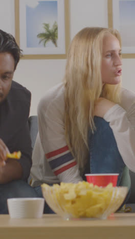 Vertical-Video-Of-Friends-Sitting-On-Sofa-At-Home-Together-Eating-Chips-And-Dips-Whilst-Watching-TV